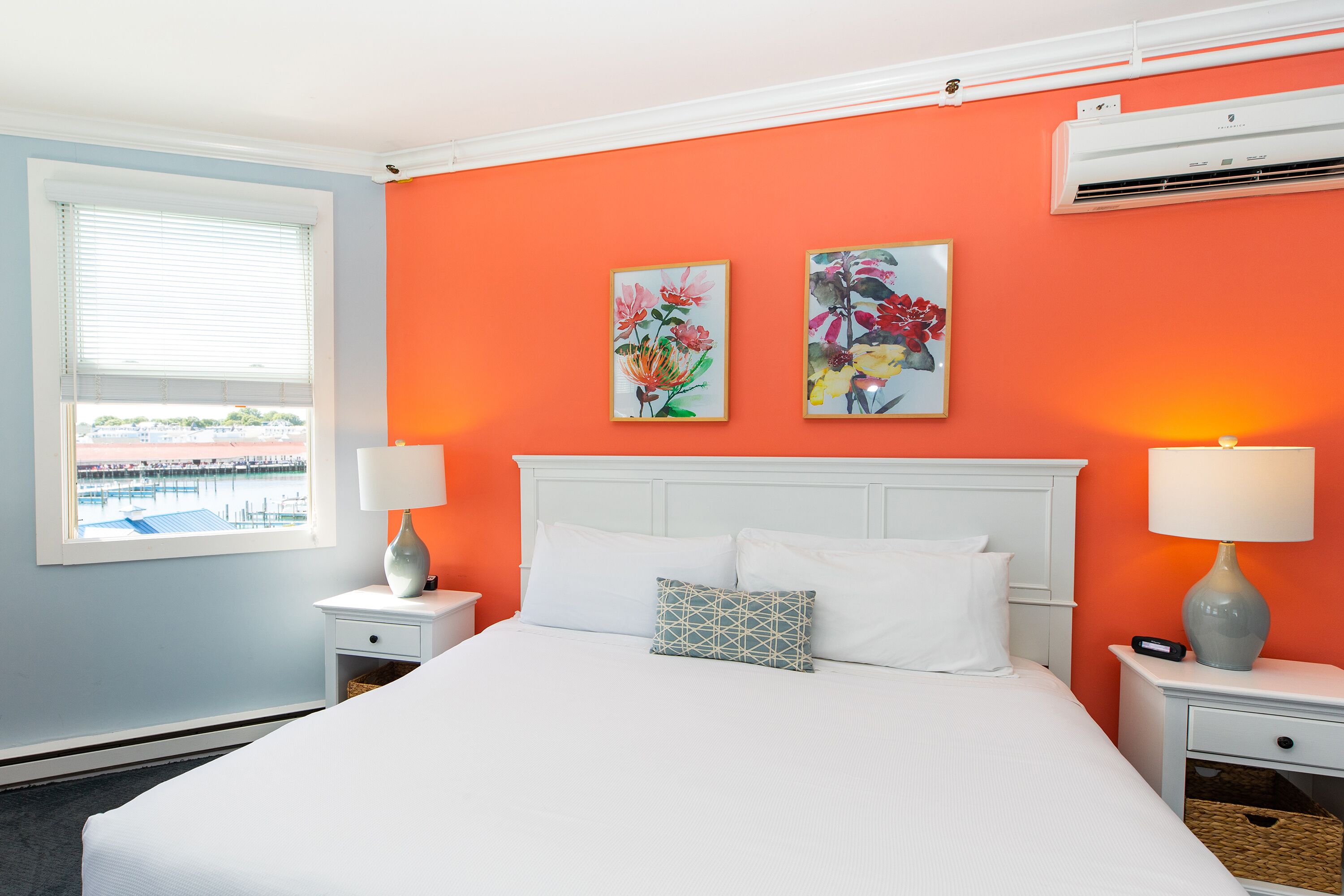 Une chambre d’hôtel comprenant un lit, deux tables de chevet, une fenêtre avec stores et deux climatiseurs fixés au mur.