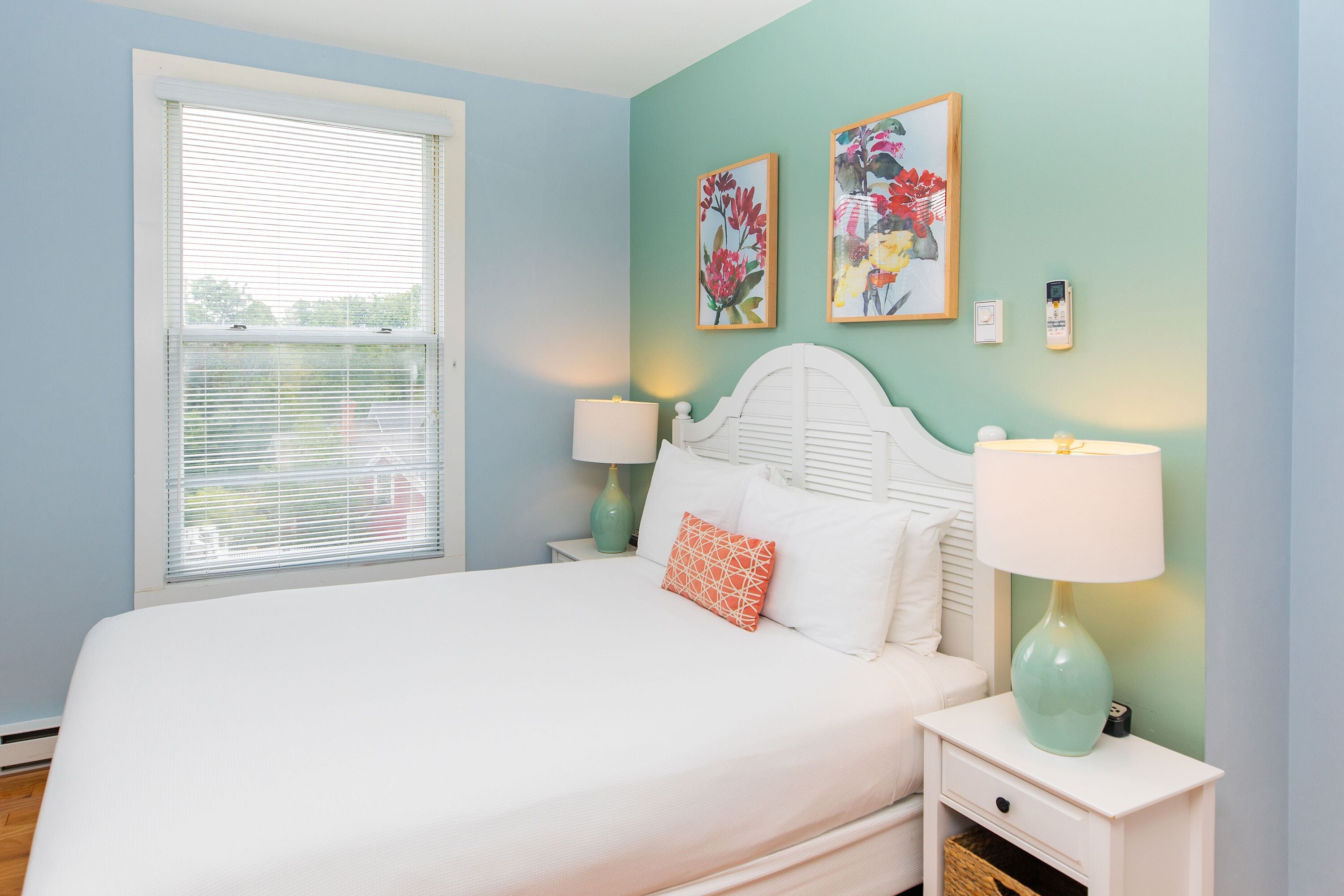 A bedroom with a bed, bedside tables, a window with blinds, and two wall-mounted paintings.