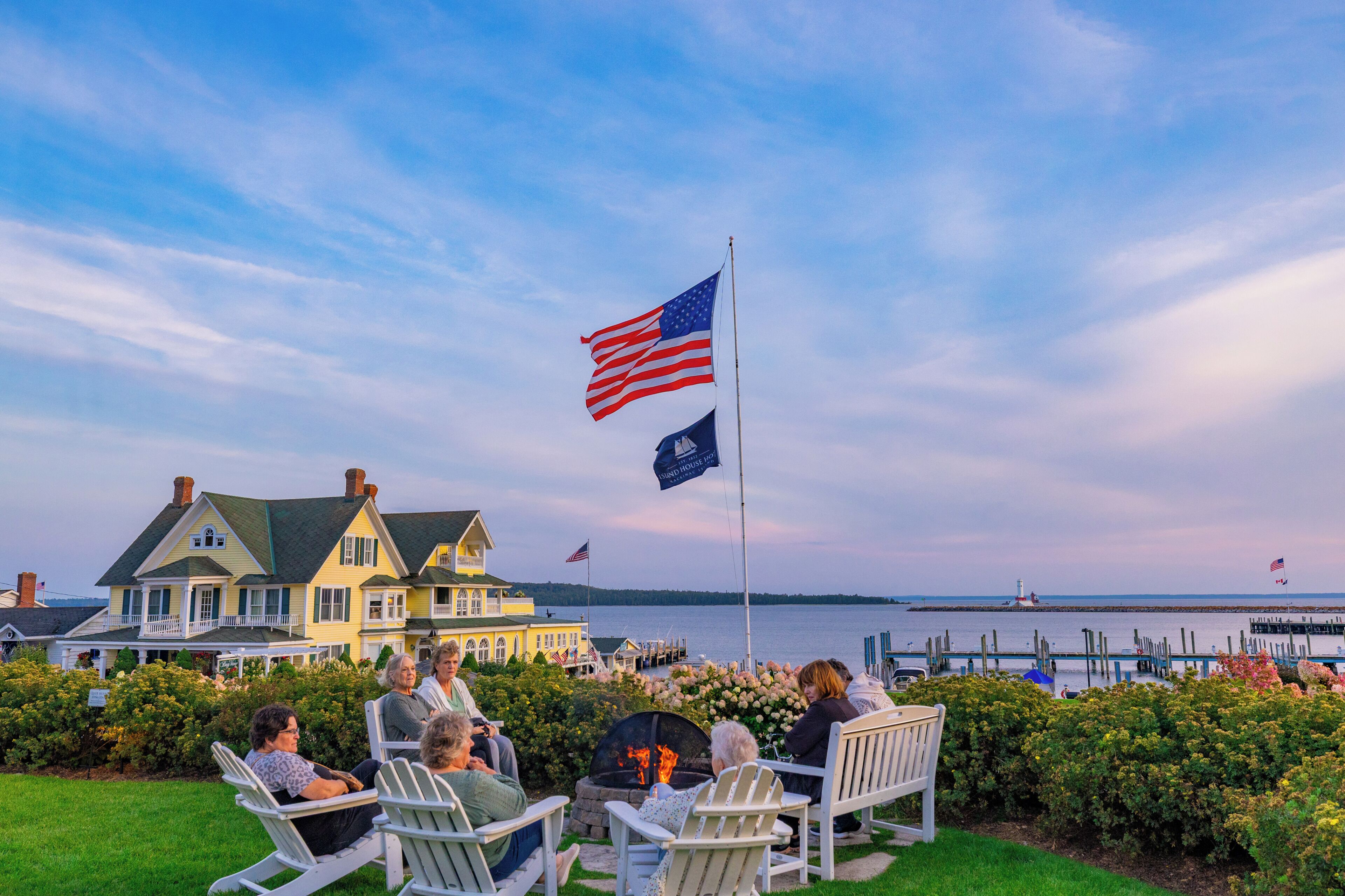 A group of people seated on Adirondack chairs by a fire pit, overlooking a body of water with a lighthouse in the distance.