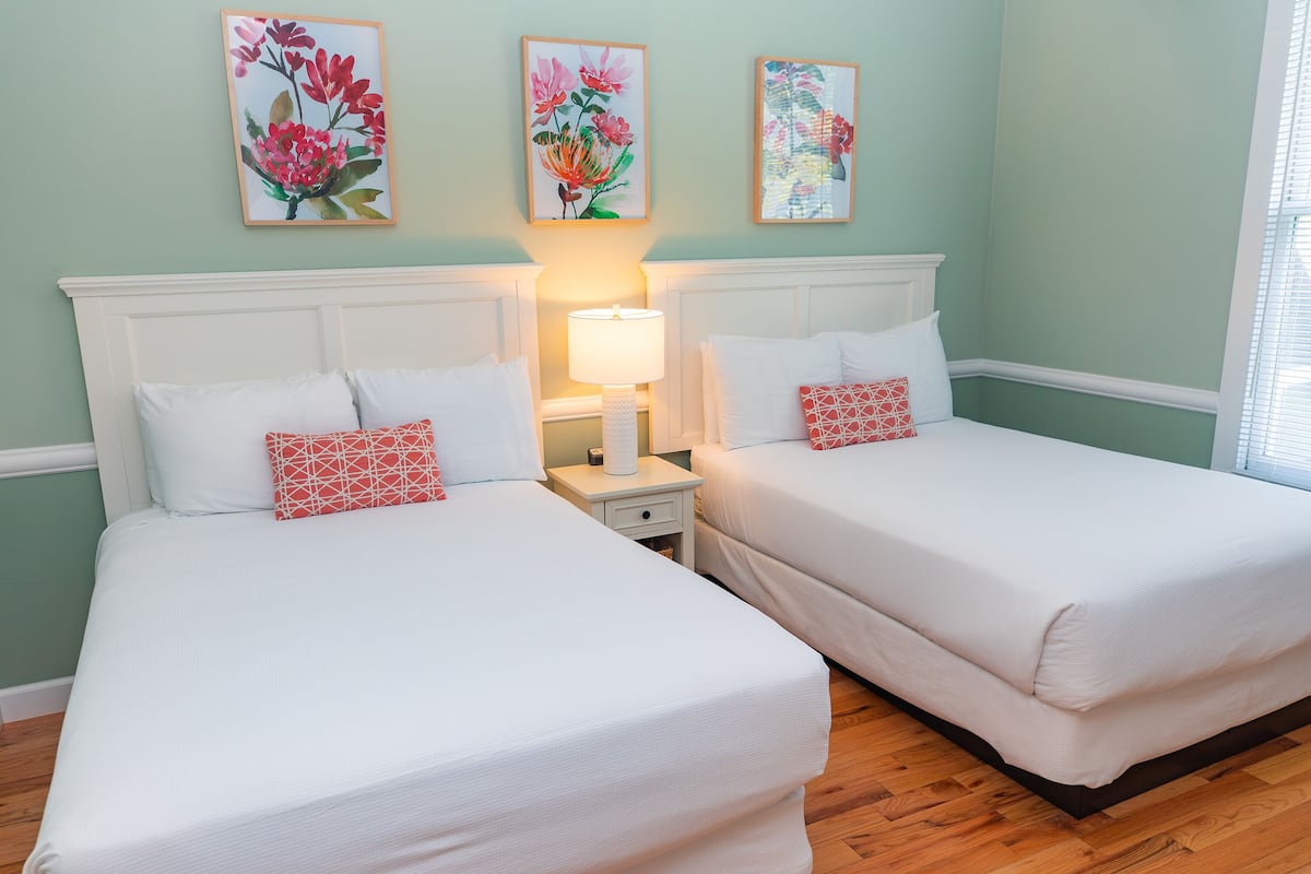 Une chambre avec deux lits, chacun doté de draps blancs et d’un coussin rouge à motifs géométriques. On y trouve deux tables de chevet avec des lampes, et les murs sont peints dans une teinte de vert clair.