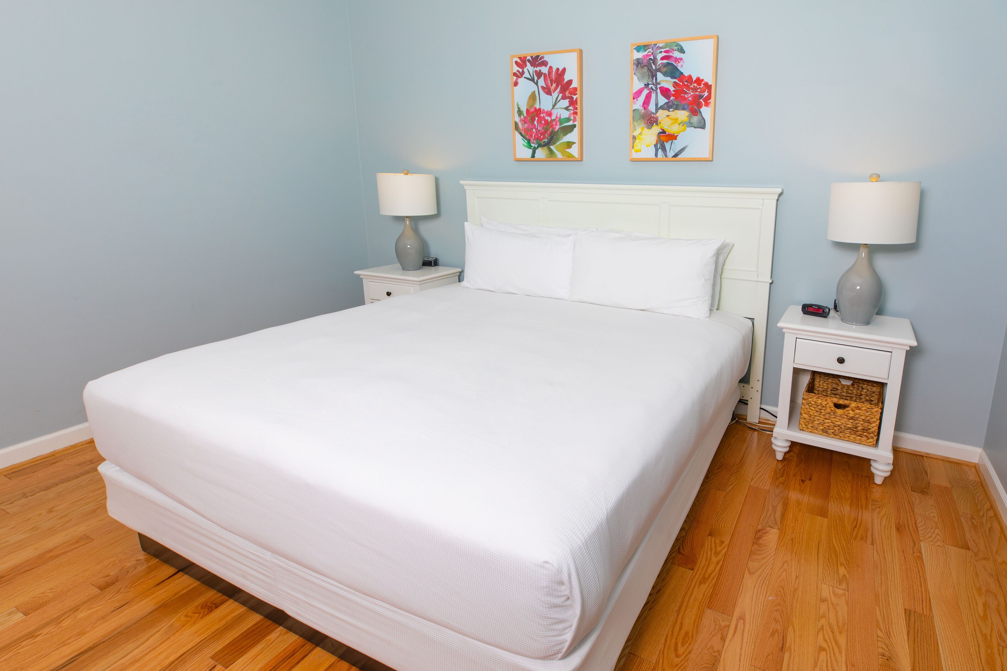 A neatly made bed with white linens in a room with light blue walls and wooden flooring.