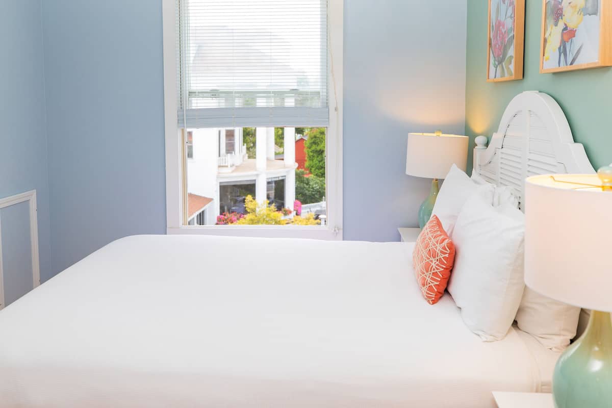 Une chambre avec un grand lit, deux lampes de chevet et une fenêtre donnant sur un balcon et des fleurs à l’extérieur.