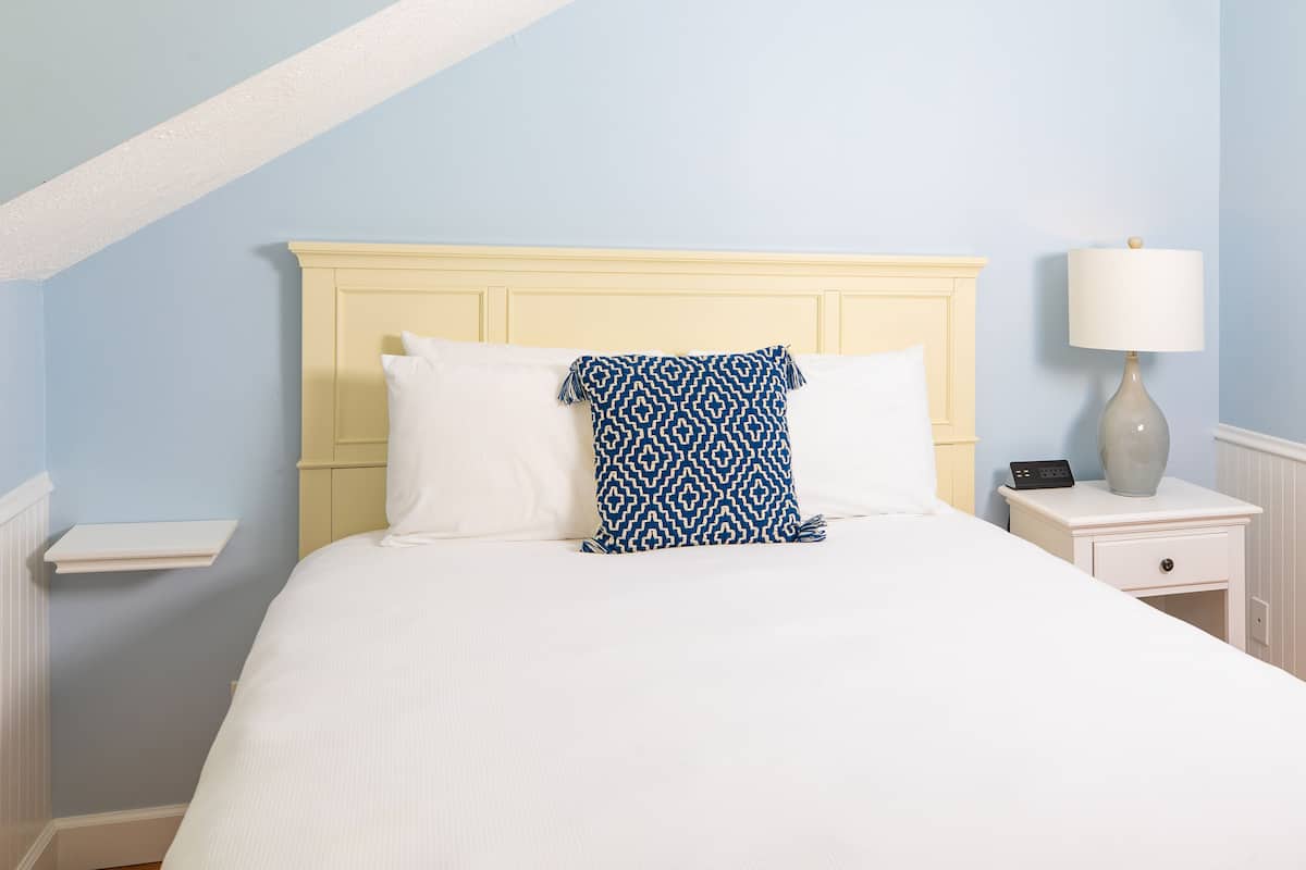 Un lit bien fait, avec des oreillers blancs et bleus à motifs, une tête de lit de couleur crème et une table de chevet sur laquelle se trouvent une lampe et un téléphone.