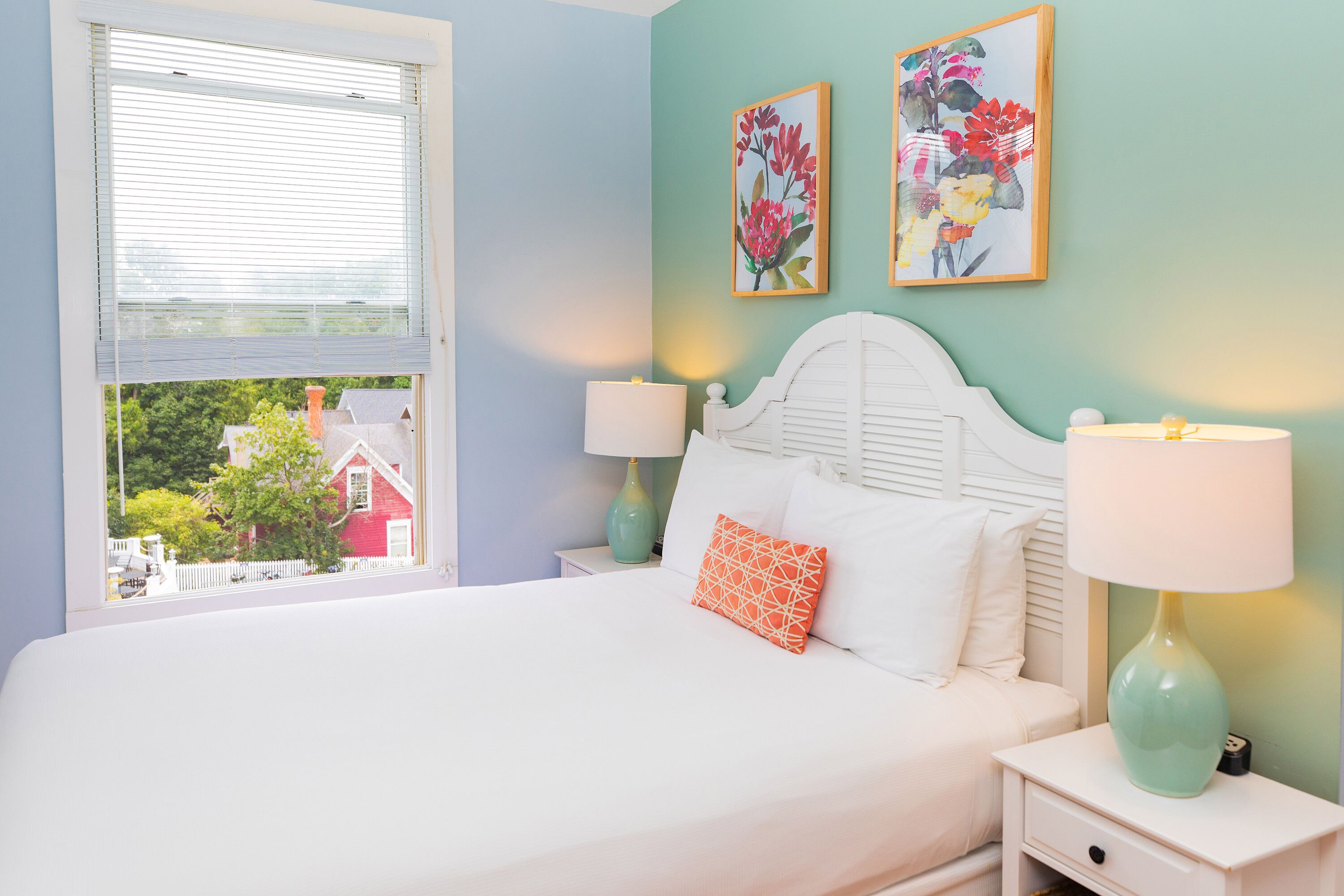 A bedroom with a bed, two bedside tables with lamps, a window with blinds, and two floral wall art pieces.