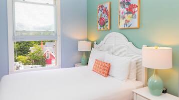 A bedroom with a bed, two bedside tables with lamps, a window with blinds, and two floral wall art pieces.