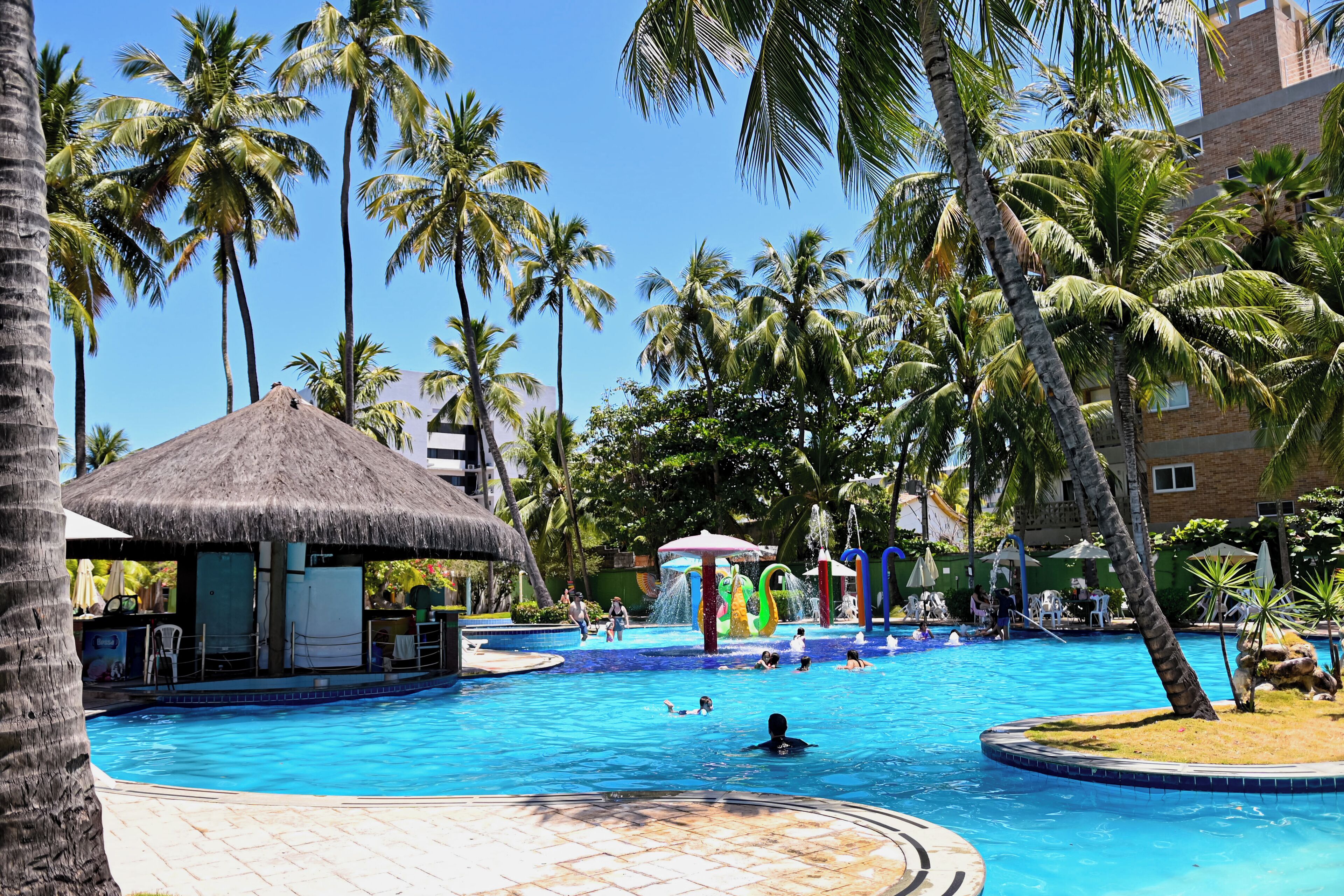 Ein Pool mit Palmen, einer strohgedeckten Hütte und einem Wasserspielbereich.