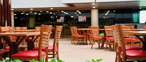 Interior de restaurante con mesas y sillas de madera, asientos acolchados rojos y una planta verde al frente.