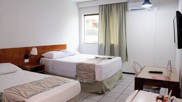 Quarto de hotel com duas camas, uma escrivaninha, uma cadeira, uma luminária, ar condicionado e uma janela com cortinas.