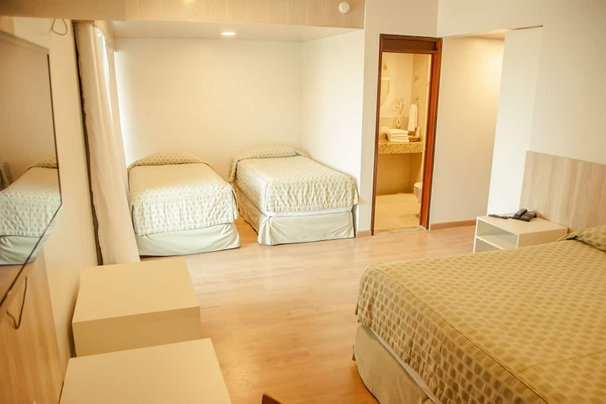 Habitación de hotel con dos camas individuales, una cama grande, un baño con espejo y una mesita con teléfono.
