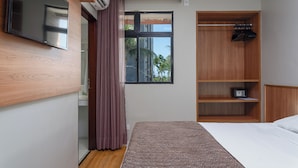 Une chambre moderne avec un lit, un téléviseur fixé au mur, une armoire en bois et une fenêtre donnant sur des palmiers.