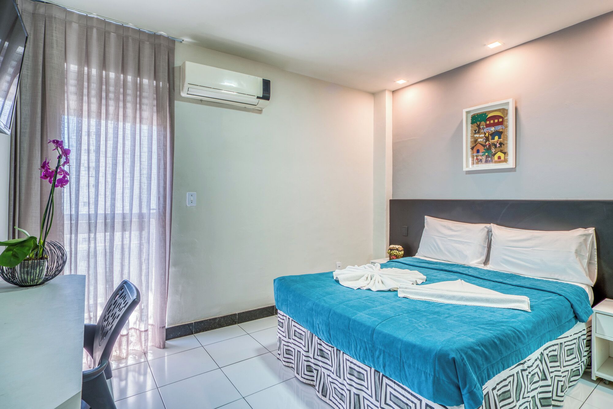 A hotel room with a bed, a desk, a chair, an air conditioner, and a window with curtains.
