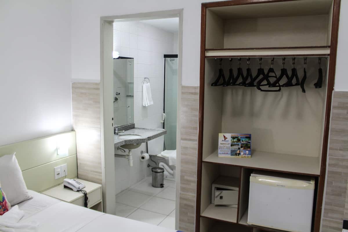 A hotel room with a bed, a bedside phone, a bathroom with a sink and mirror, and a coat rack with hanging items.