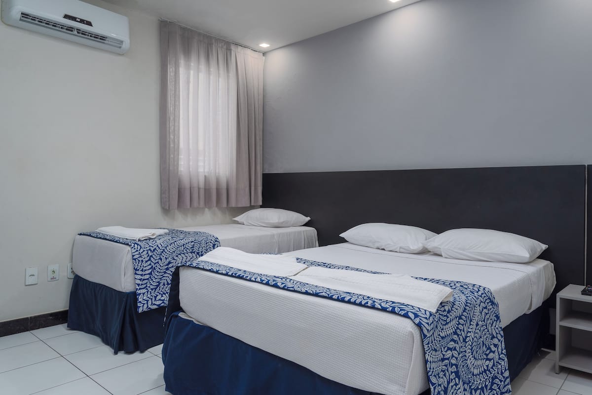 A hotel room with two single beds, a large window with sheer curtains, a ceiling fan, and a small bedside table.