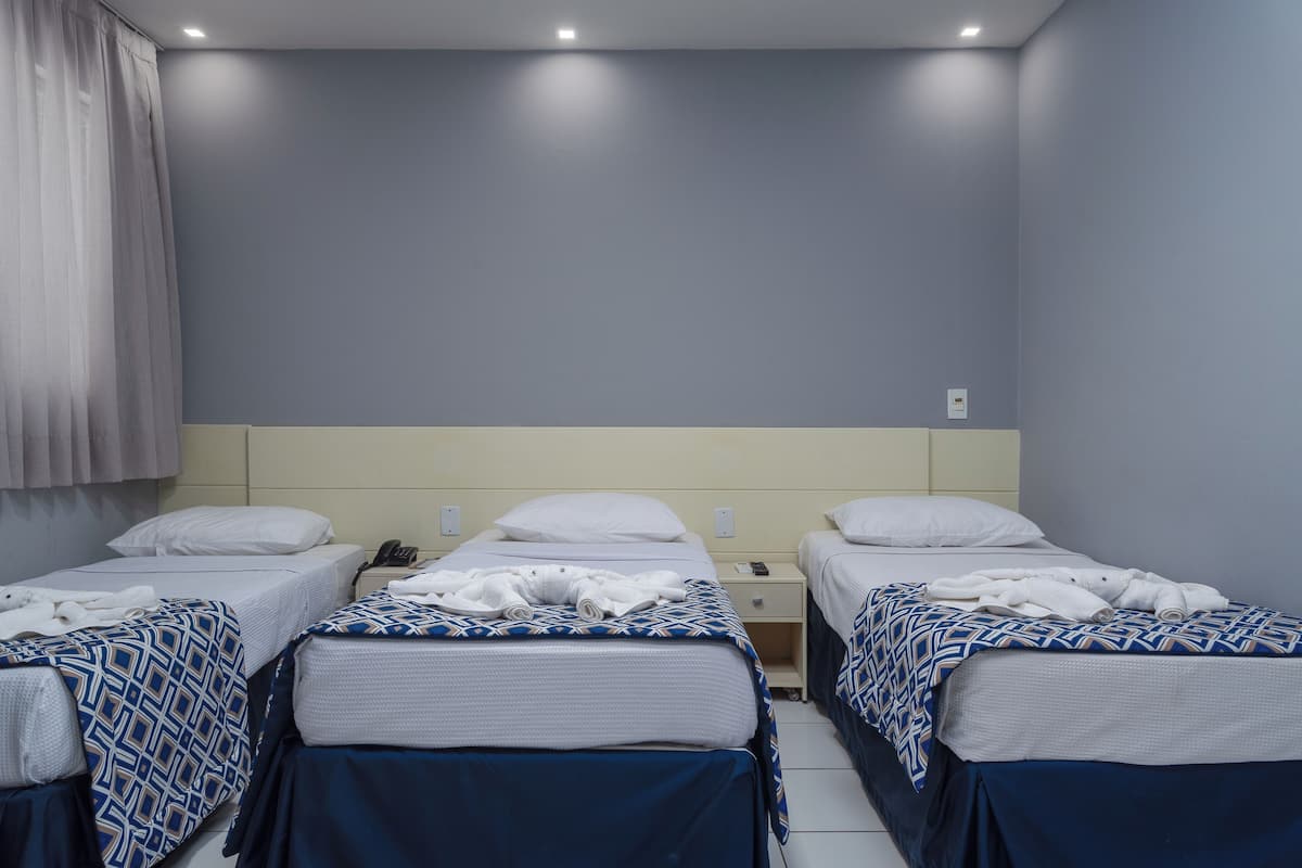 A hotel room with three single beds, each with a patterned blanket and white pillows. There is a bedside table with a phone and a lamp.