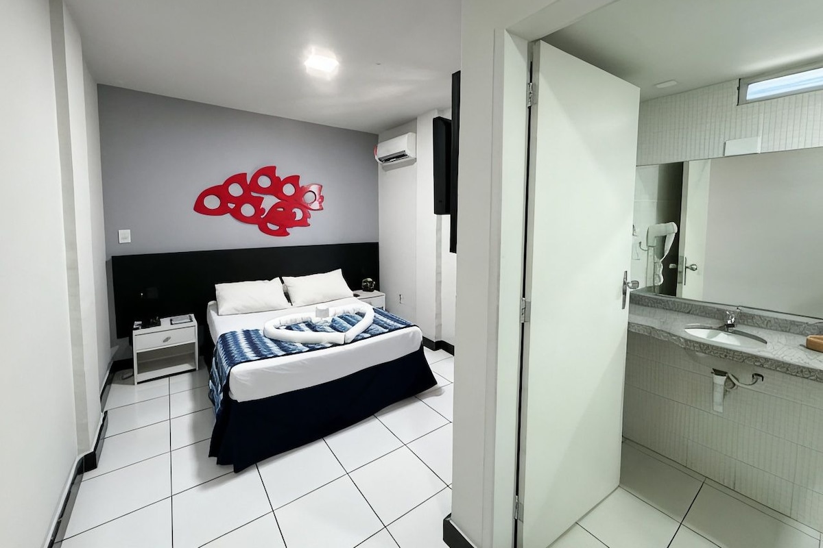 Habitación de hotel moderna con cama, mesita de noche, baño con lavabo y espejo, y una decoración abstracta roja en la pared.