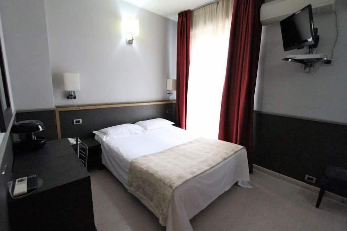 A hotel room with a bed, a desk with a telephone, a television mounted on the wall, and a window with curtains.