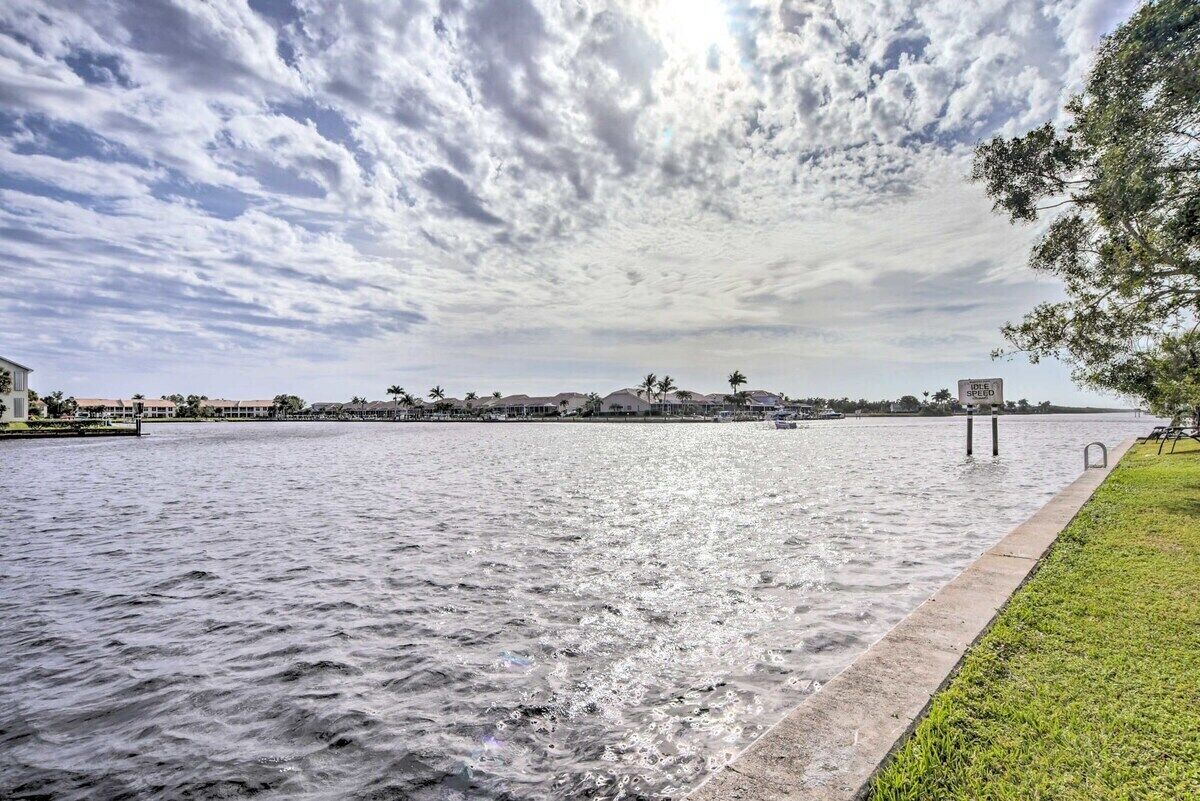 A waterfront view with a calm river, a grassy area, and a distant building with palm trees.