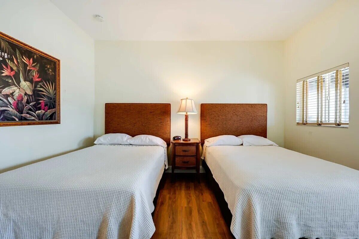 A hotel room with two single beds, a nightstand with a lamp, a painting on the wall, and a window with blinds.