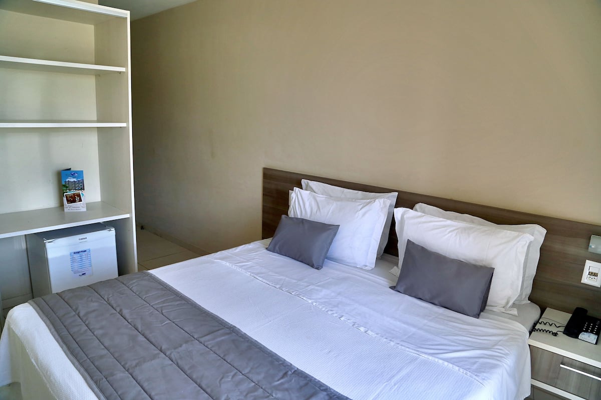 A neatly made bed with white and gray bedding, a small refrigerator, and a bedside table with a phone and a lamp.