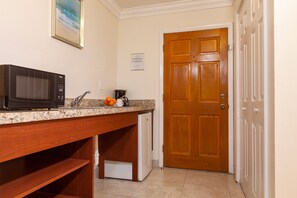 Una cocina con microondas, lavamanos y refrigerador. Se ve una puerta de madera y una puerta blanca al fondo.