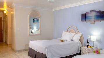 A hotel room with two beds, a large mirror, a wall art piece, and a bedside table with a wine bottle and fruit.