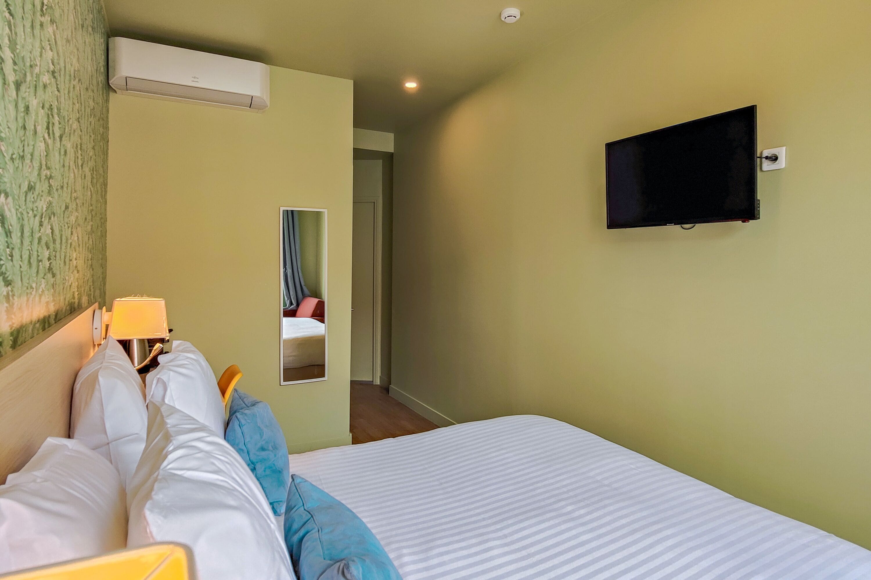 Ein Hotelzimmer mit einem Bett, einem an der Wand befestigten Fernseher, einer Klimaanlage und einem Spiegel, der einen Teil des Raumes widerspiegelt.