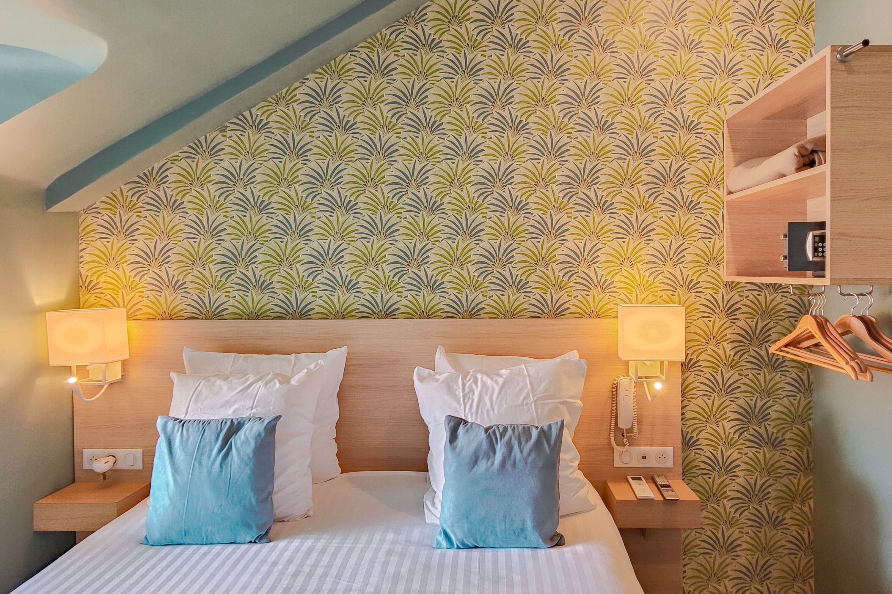 Una habitación de hotel con cama, dos mesitas de noche, un tocador, un estante en la pared y un perchero.