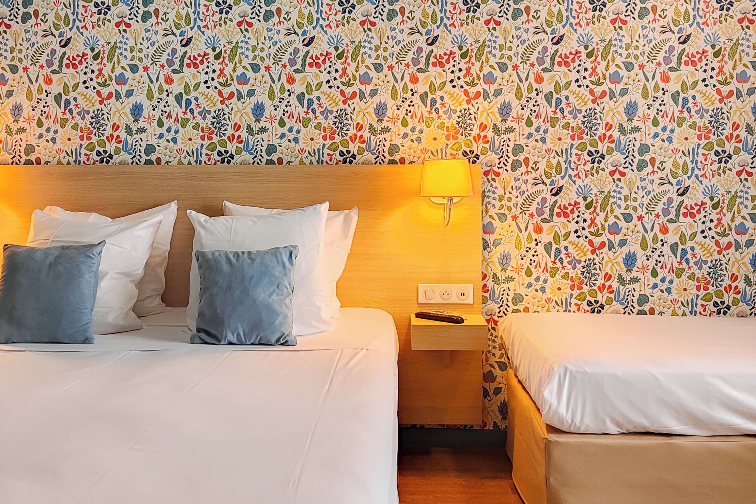 A hotel room with two beds, a wooden headboard, a nightstand, and a wall with a floral pattern.