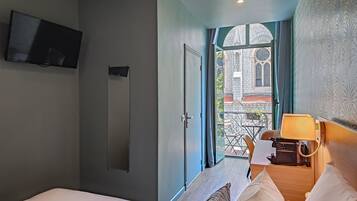 Habitación de hotel con cama, escritorio con lámpara, televisor y vista a un edificio por una puerta de vidrio.