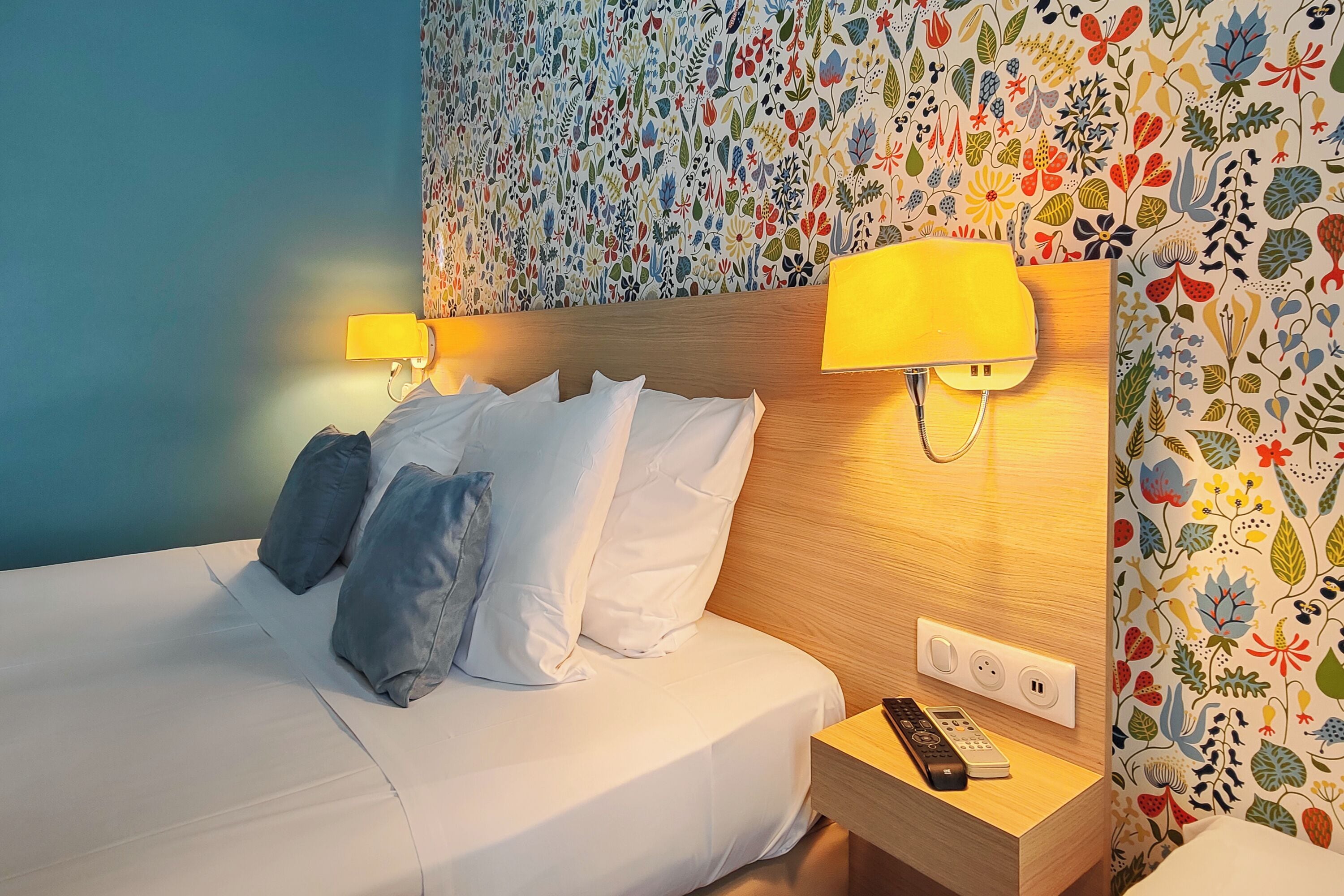 A hotel room with a bed, two pillows, a nightstand with a remote control, and a wall with a floral pattern.