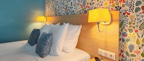 A hotel room with a bed, two pillows, a nightstand with a remote control, and a wall with a floral pattern.