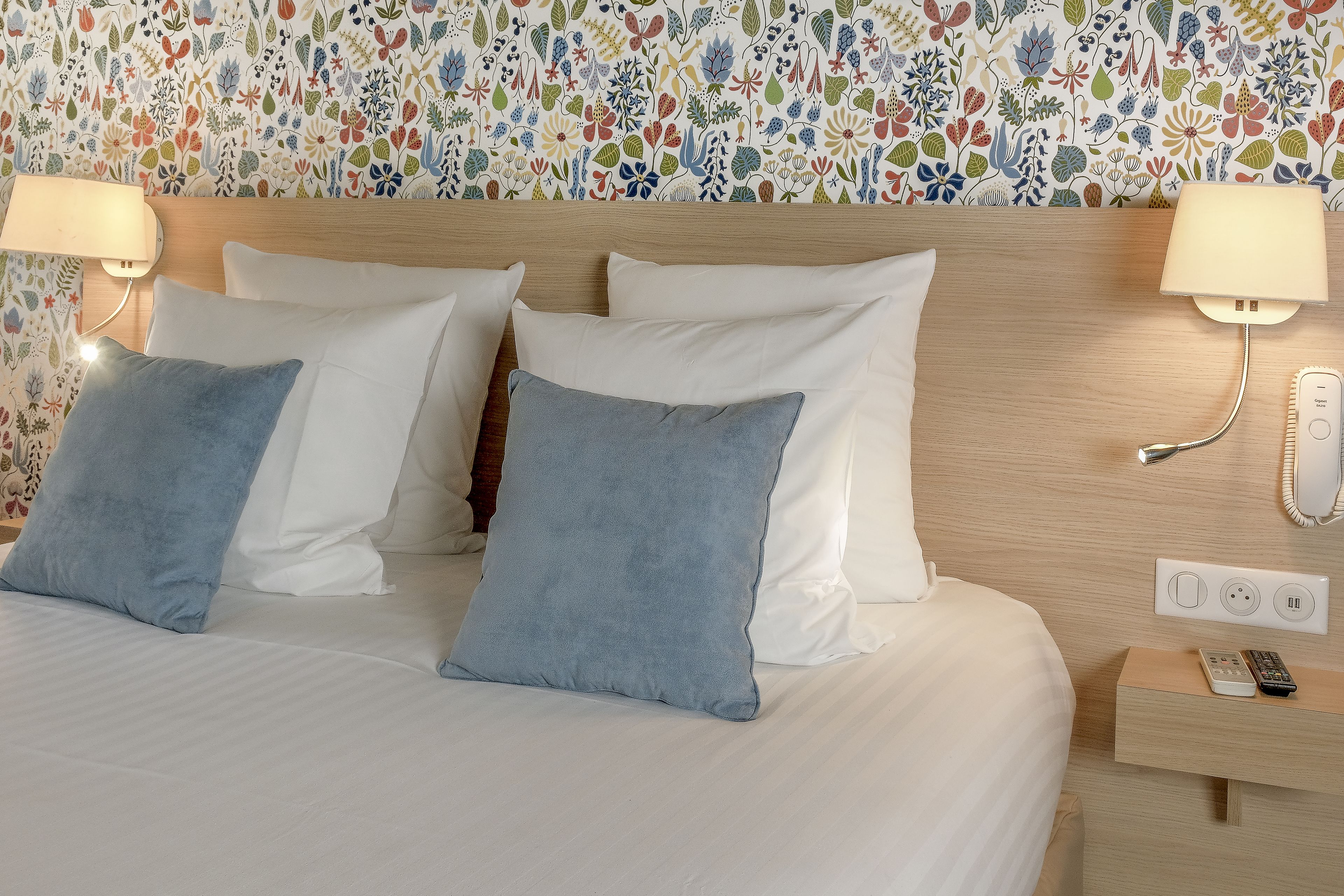 Hotelkamer met een bed, twee bedlampjes, een nachtkastje met telefoon en afstandsbediening, en een wand met bloemenprint.