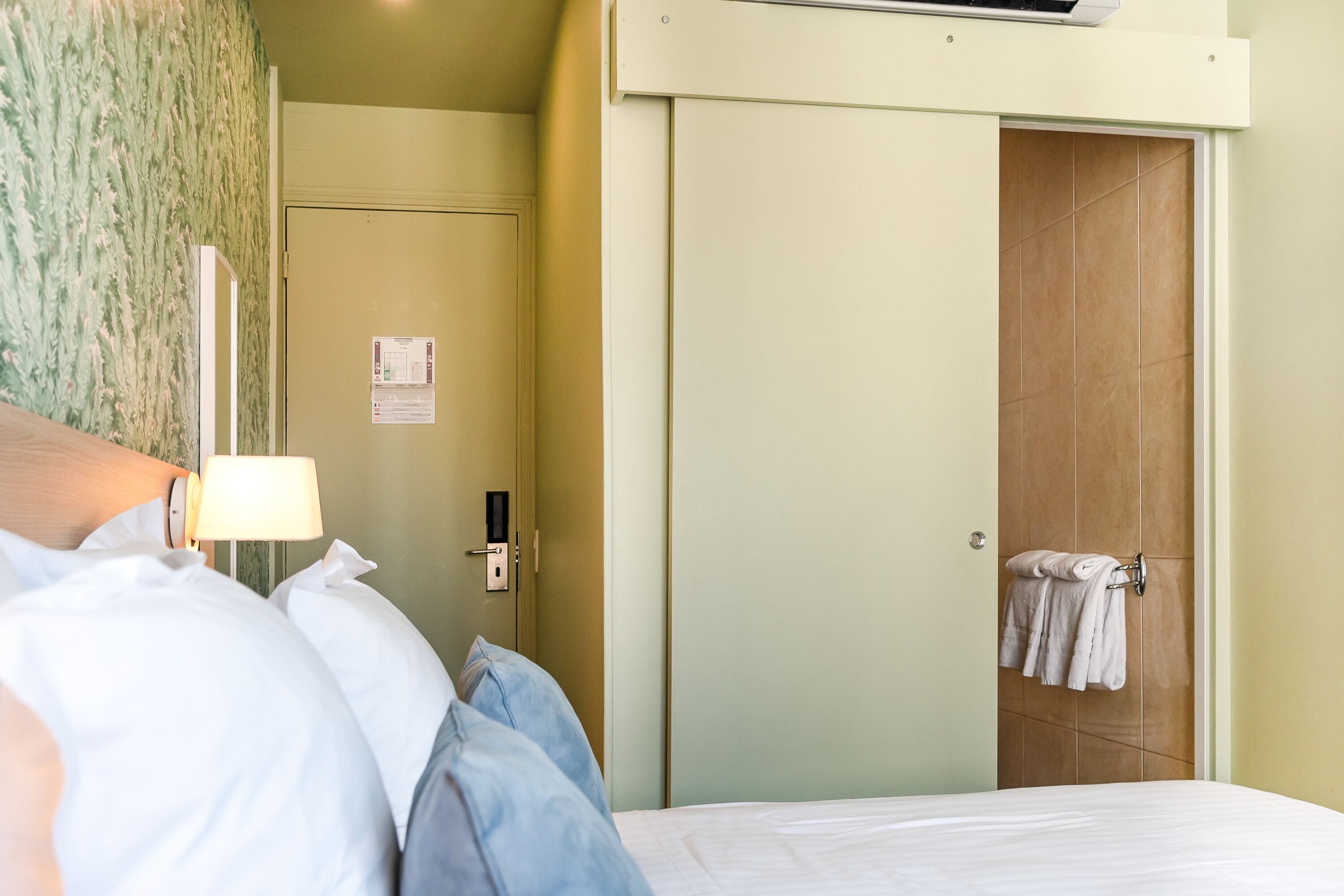 A hotel room with a bed, a nightstand, a lamp, a door, and a towel rack.