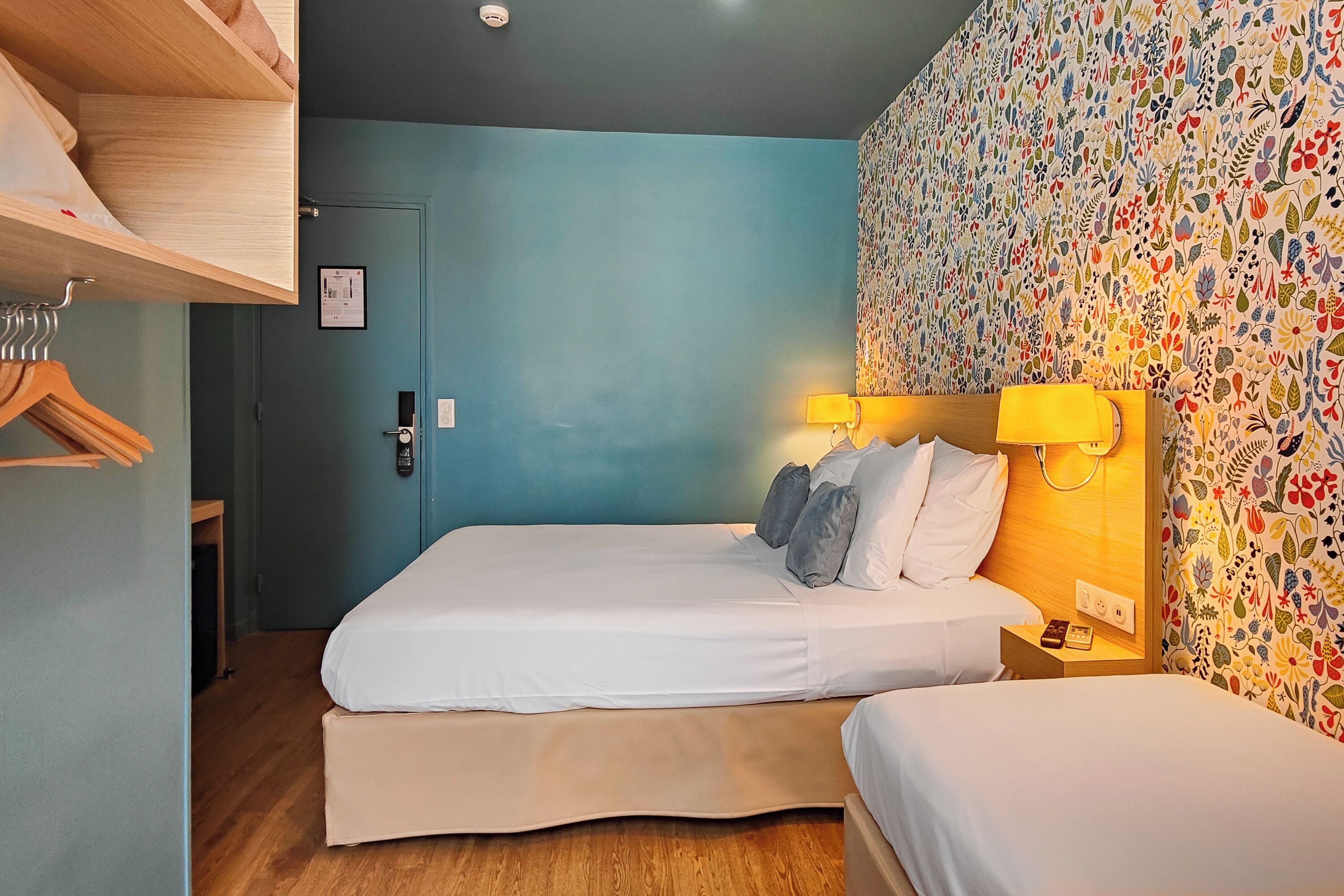 Ett hotellrum med två sängar, en garderob i trä, en vägg med ett blommönster och en dörr med lås.