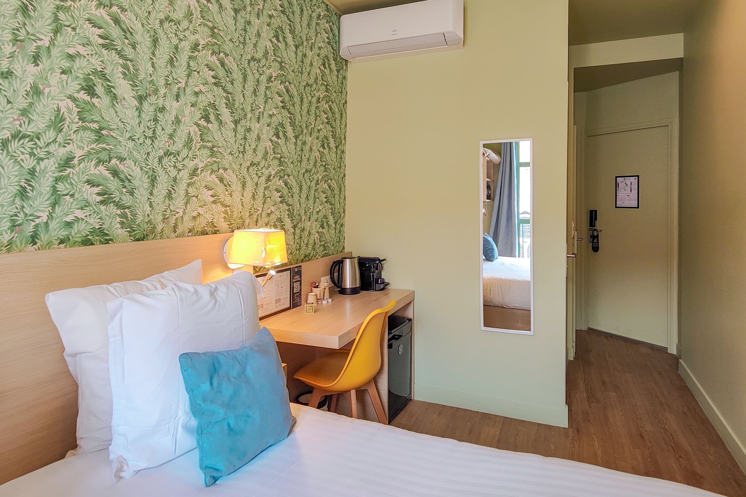 Ein Hotelzimmer mit einem Bett, einem Schreibtisch, einem Stuhl, einer Lampe, einer Kaffeemaschine und einer Klimaanlage.