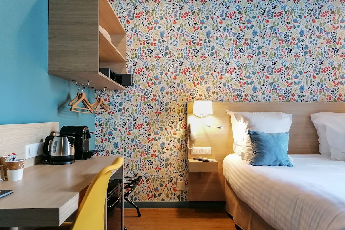 A hotel room with a bed, bedside table, lamp, and a desk with a kettle and a cup.