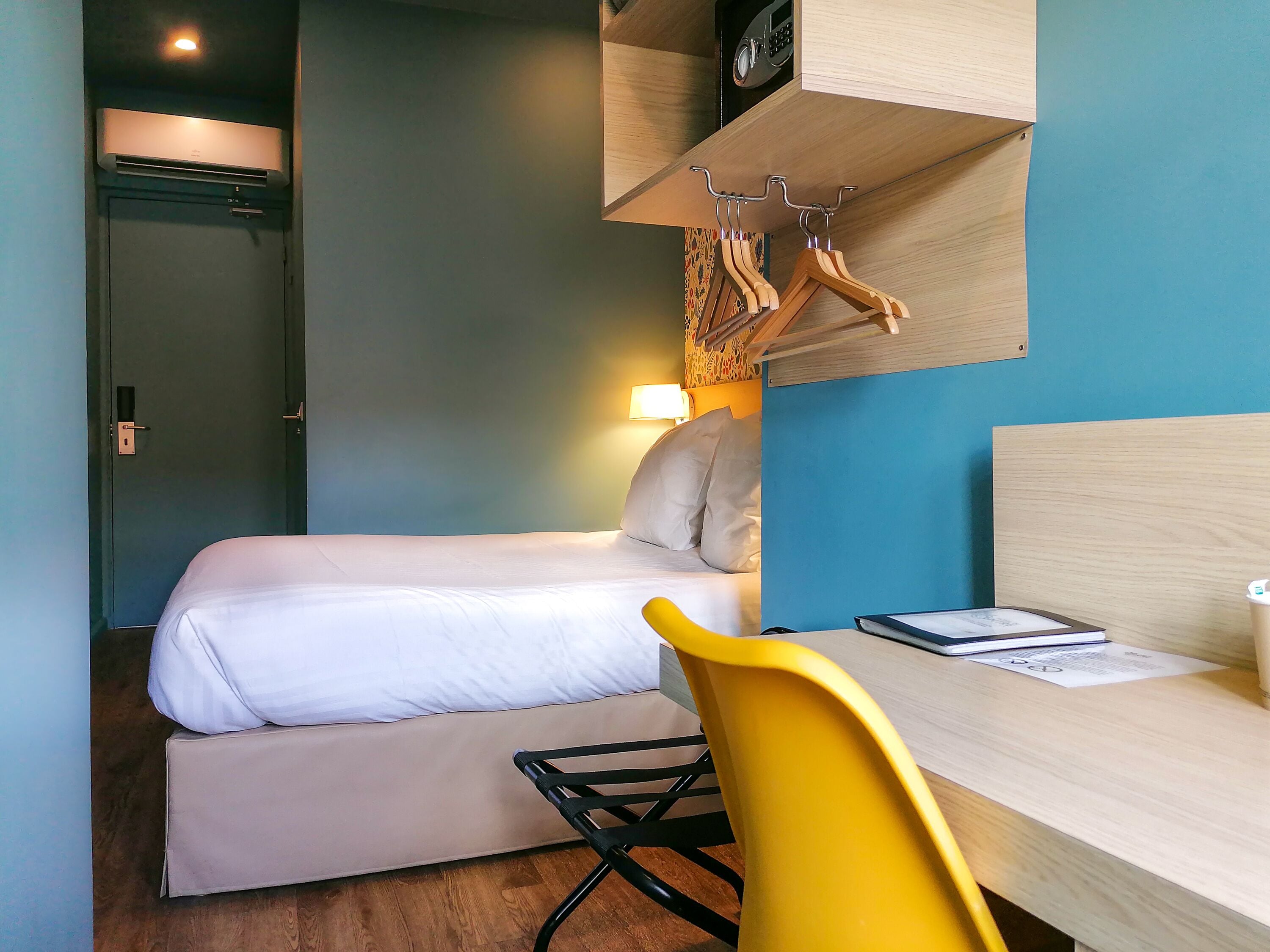 Habitación de hotel pequeña con cama, escritorio, silla y perchero en la pared.