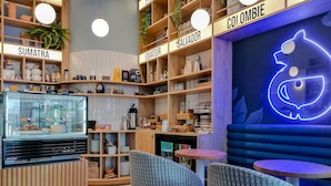 A cozy cafe with wooden shelves labeled with coffee origins like 'Sumatra' and 'Colombie', a neon pig sign, and comfortable seating. - Hotel Du Centre, un hotel AMMI (Nice)