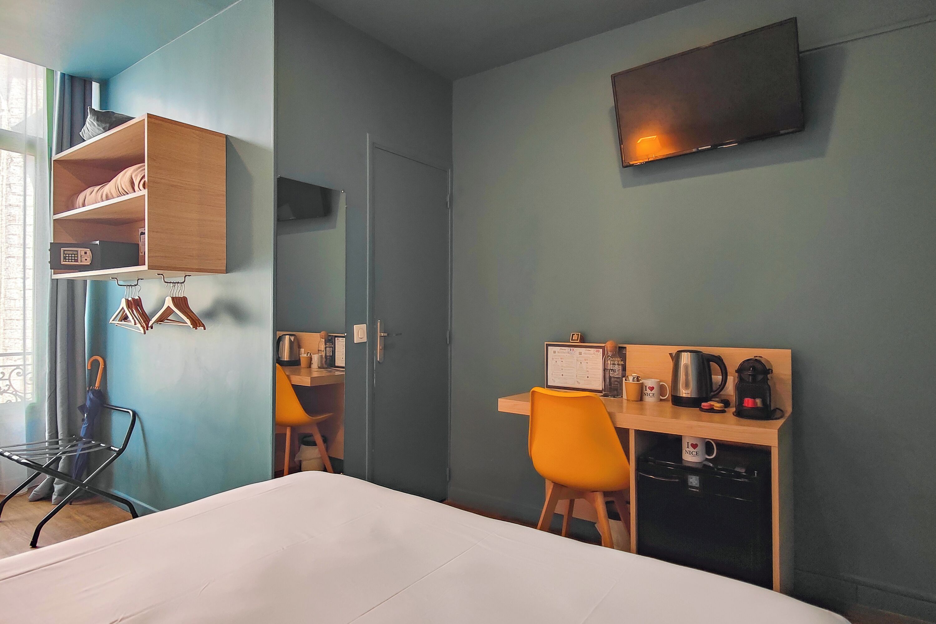 Habitación de hotel con cama, escritorio con silla, televisor, estante con un gato y ventana con cortinas.