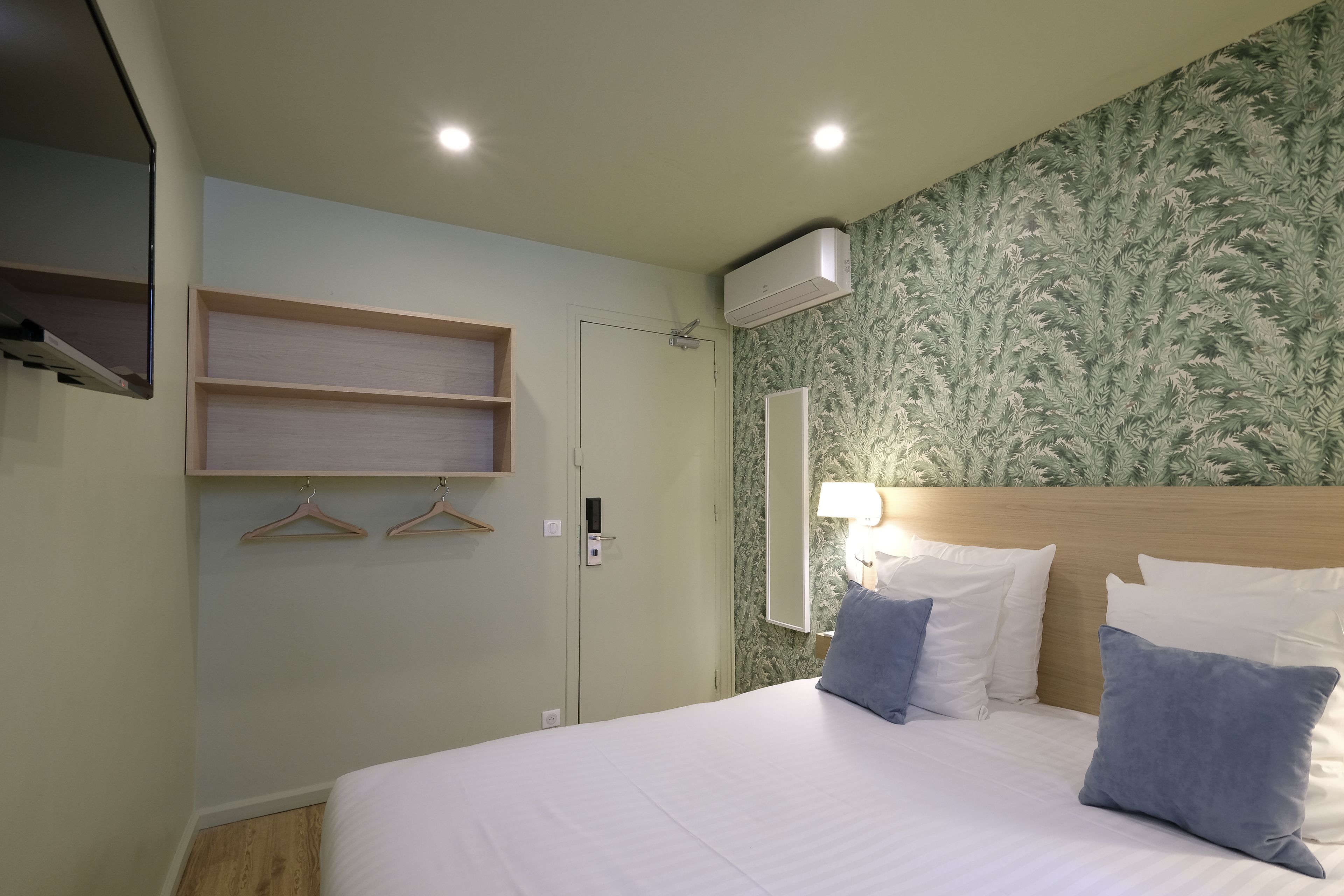 A hotel room with a bed, a wall-mounted air conditioner, a wooden shelf, and a door.