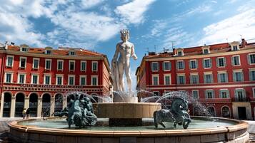 A fountain with a statue of a man in a crown, surrounded by water jets, in front of a red building with multiple windows and balconies.