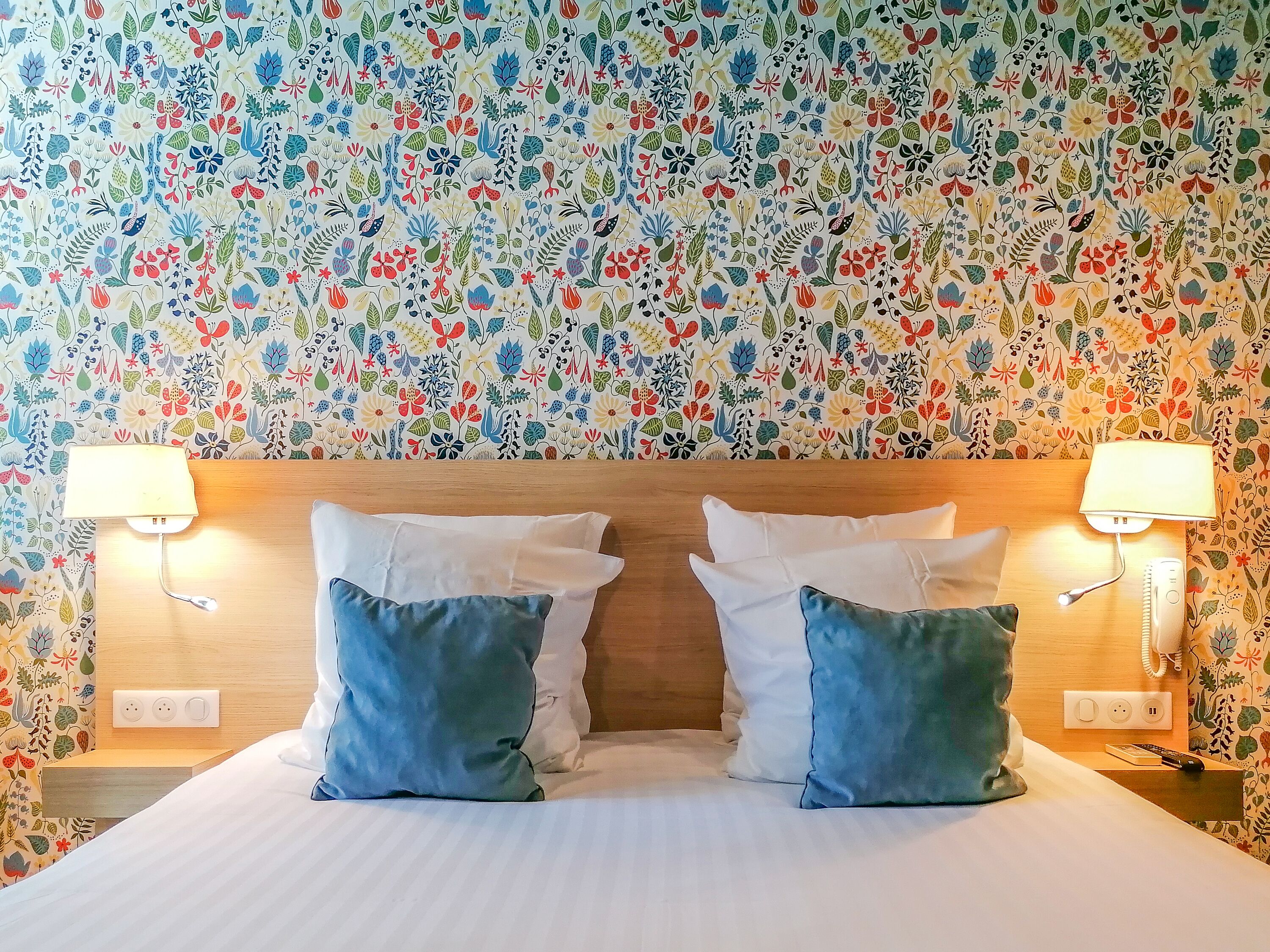 Ein Hotelzimmer mit floralem Wandpapier, einem hölzernen Kopfteil, zwei Nachttischlampen und einem Bett mit weißen und blauen Kissen.