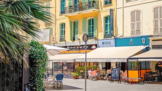 A hotel with multiple balconies, outdoor seating, and a cafe in the foreground. - Hotel Du Centre, un hotel AMMI (Nice)