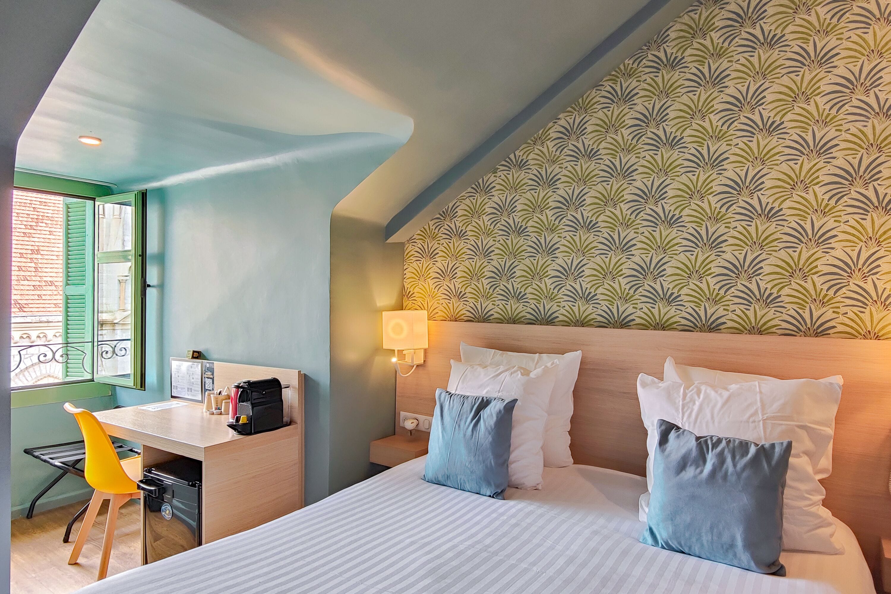 A hotel room with a bed, a desk, a chair, a nightstand, and a wall with a leafy pattern.