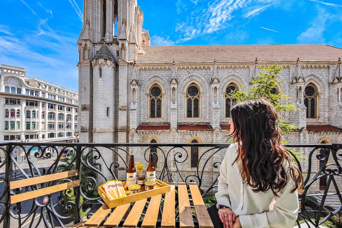 A woman sitting at a wooden table with a tray of food and drinks, overlooking a historic building with a prominent tower and arched windows.