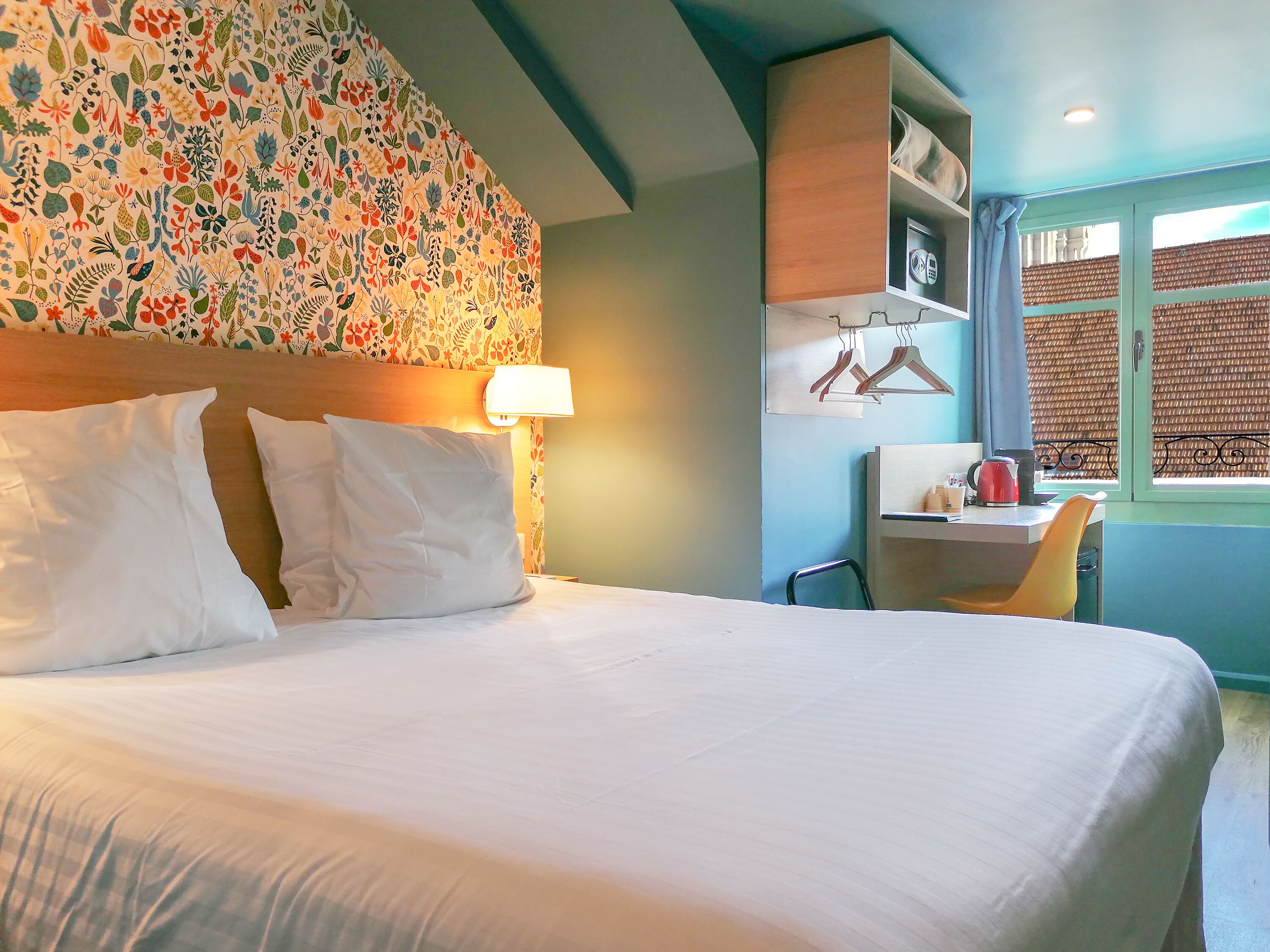 Une chambre d’hôtel avec un grand lit, un bureau avec une chaise, une lampe et une fenêtre donnant sur un immeuble.