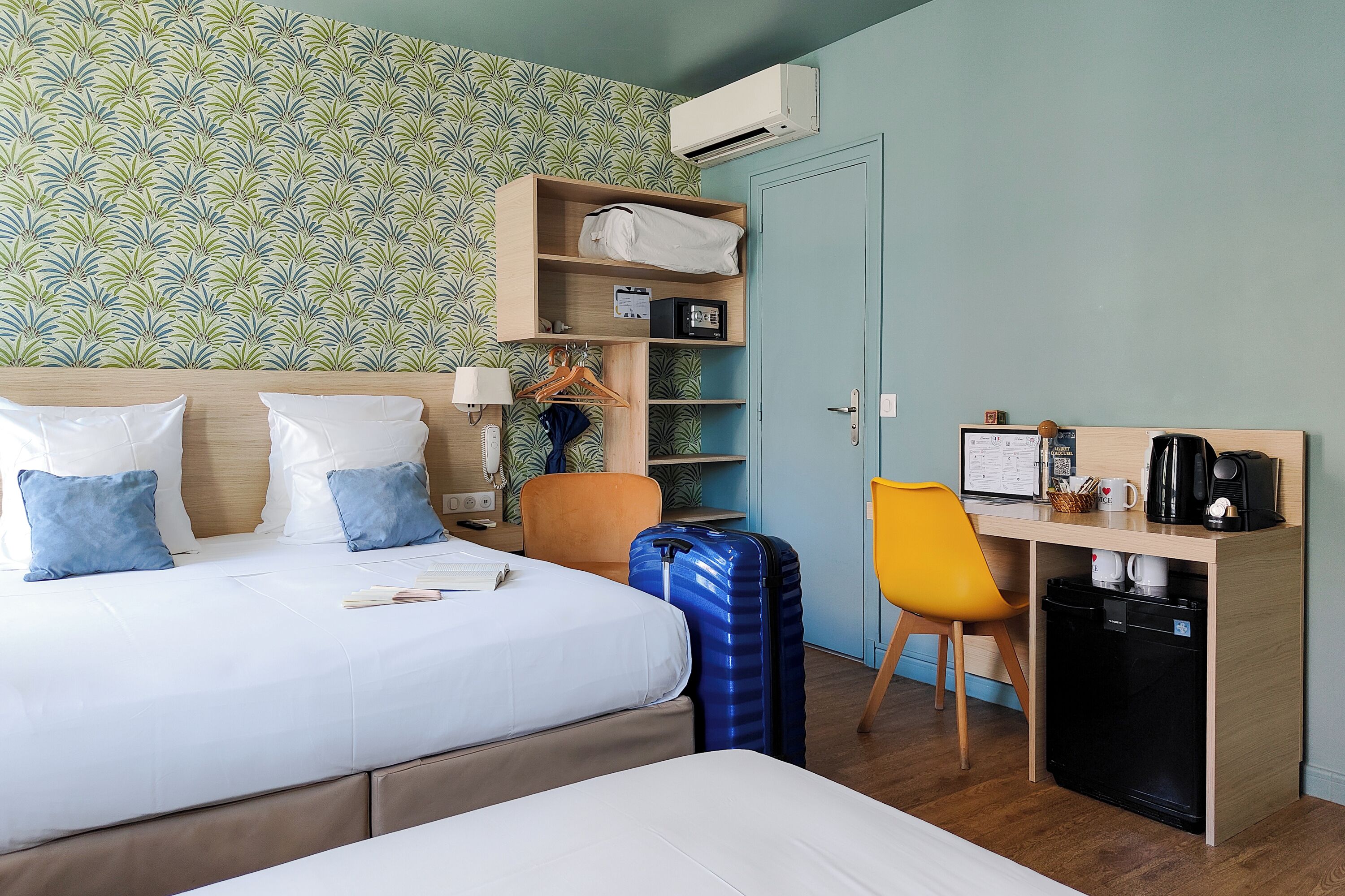 Ett hotellrum med en säng, ett skrivbord med en dator, en stol, en resväska och ett litet kylskåp.