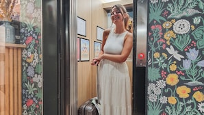 A woman in a white dress standing in an elevator with floral wallpaper and framed certificates on the wall. - Hotel Du Centre, un hotel AMMI (Nice)