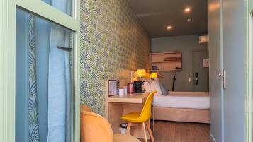 A compact hotel room with a bed, a desk with a chair, a small table with a lamp, and a wardrobe.