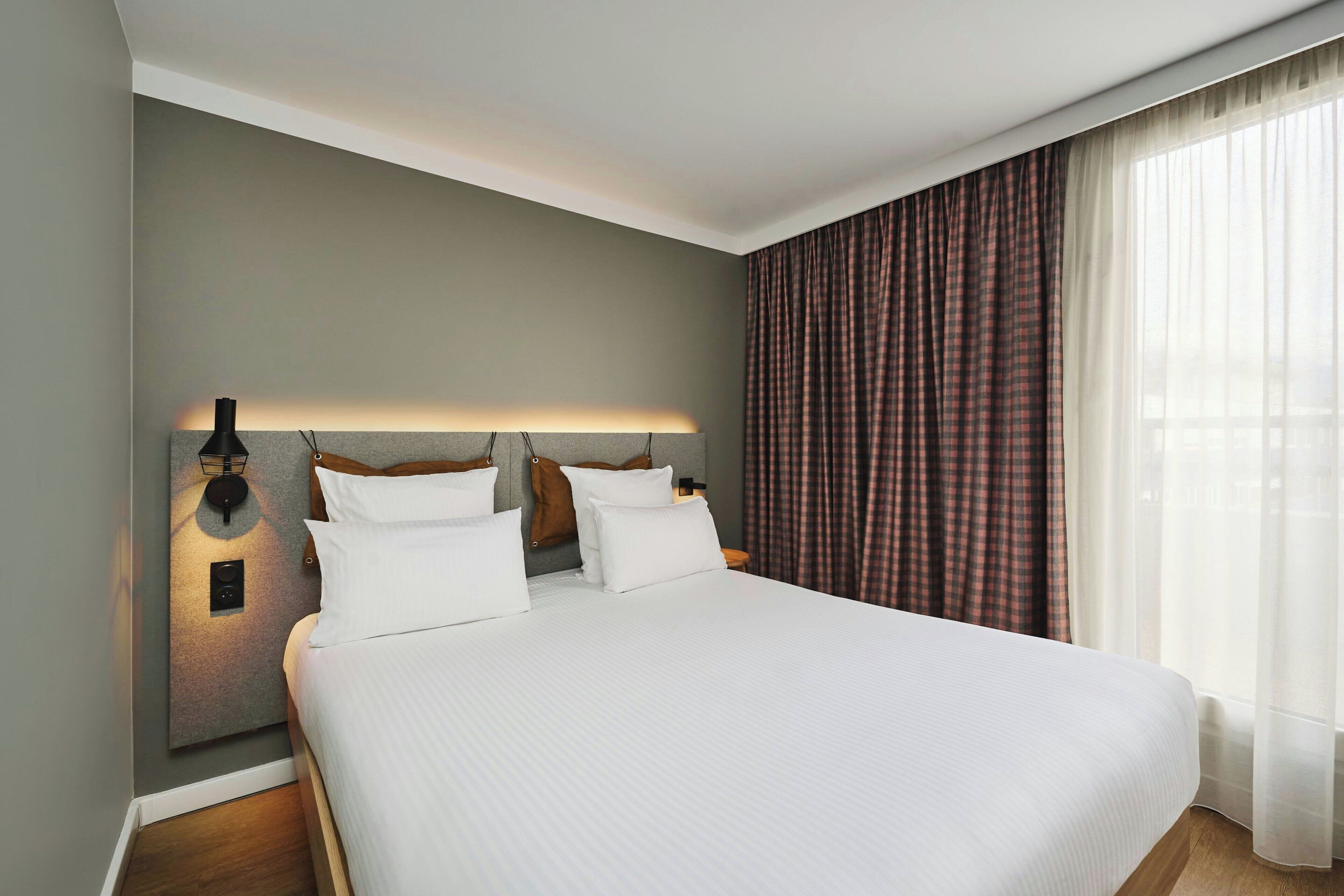 A hotel room with a large bed, a nightstand, a wall-mounted lamp, and a window with curtains.